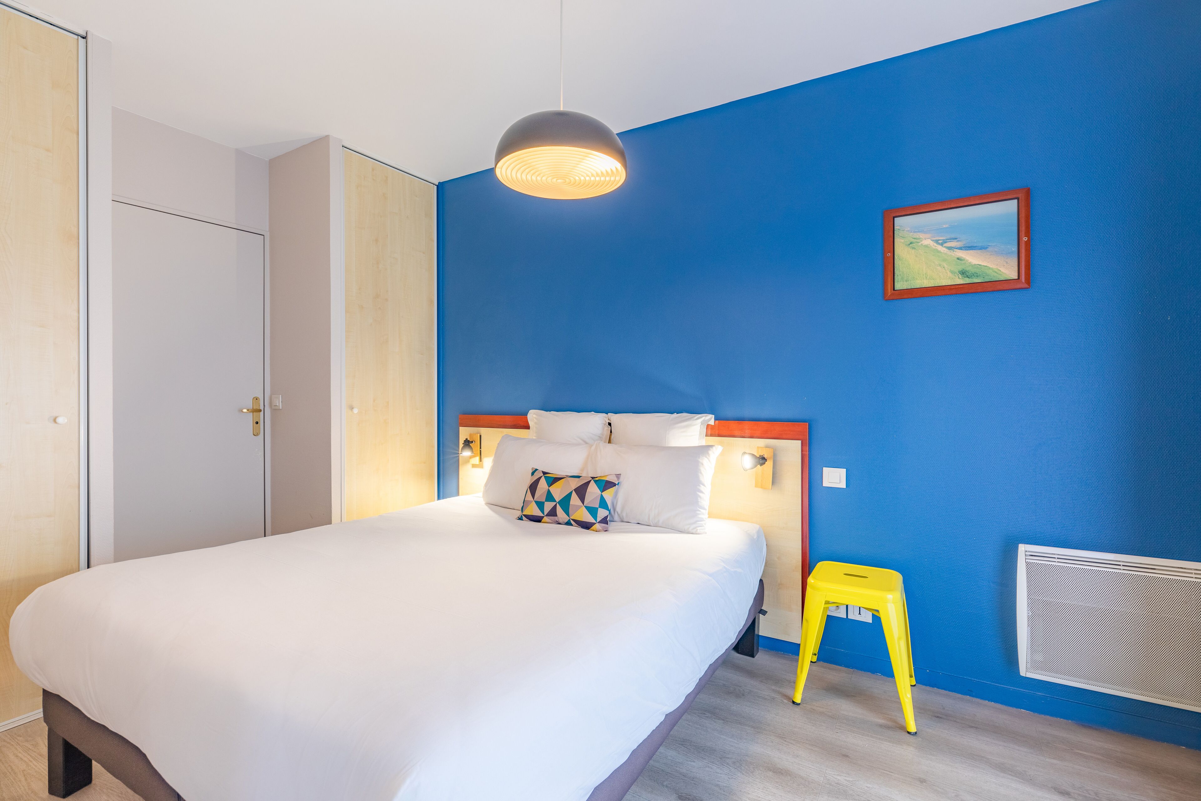 A bedroom with a bed, a nightstand, a yellow stool, a picture on the wall, and a window.