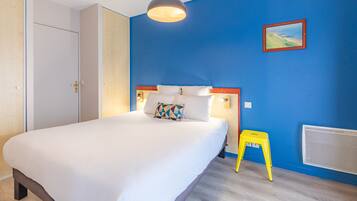 Une chambre à coucher avec un lit, une table de chevet, un tabouret jaune, un tableau accroché au mur et une fenêtre.