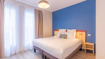 A neatly made bed with white bedding and a geometric pillow, a bedside table, and a wall-mounted light fixture.
