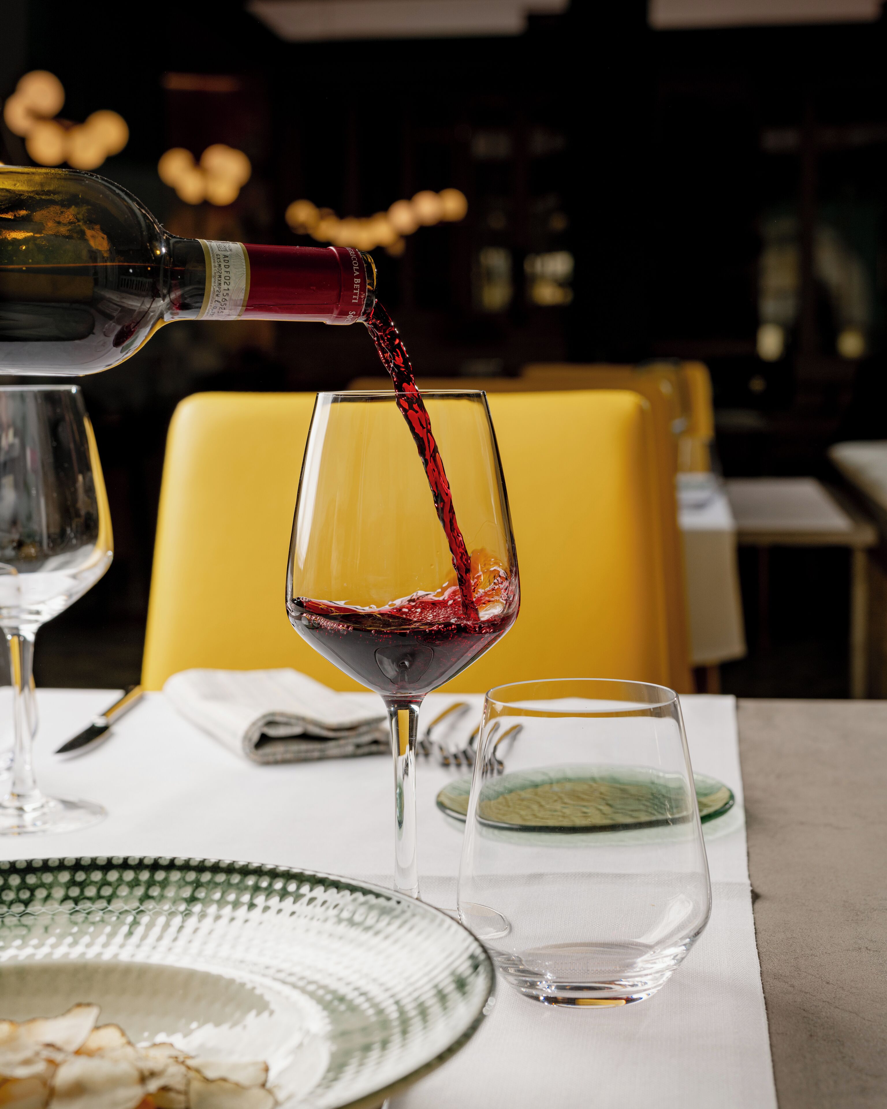 A dining table with a wine glass being filled with red wine, a glass of water, and a green plate.