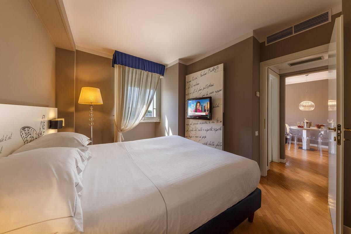 A hotel room with a large bed, a bedside lamp, a TV, and a view of the dining area.