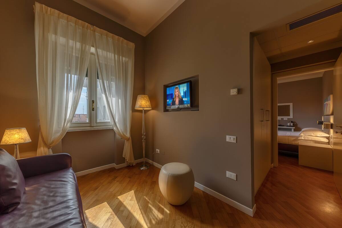 A modern hotel room with a wooden floor, a large window with sheer curtains, a flat-screen TV mounted on the wall, a round ottoman, and a bed with a headboard.