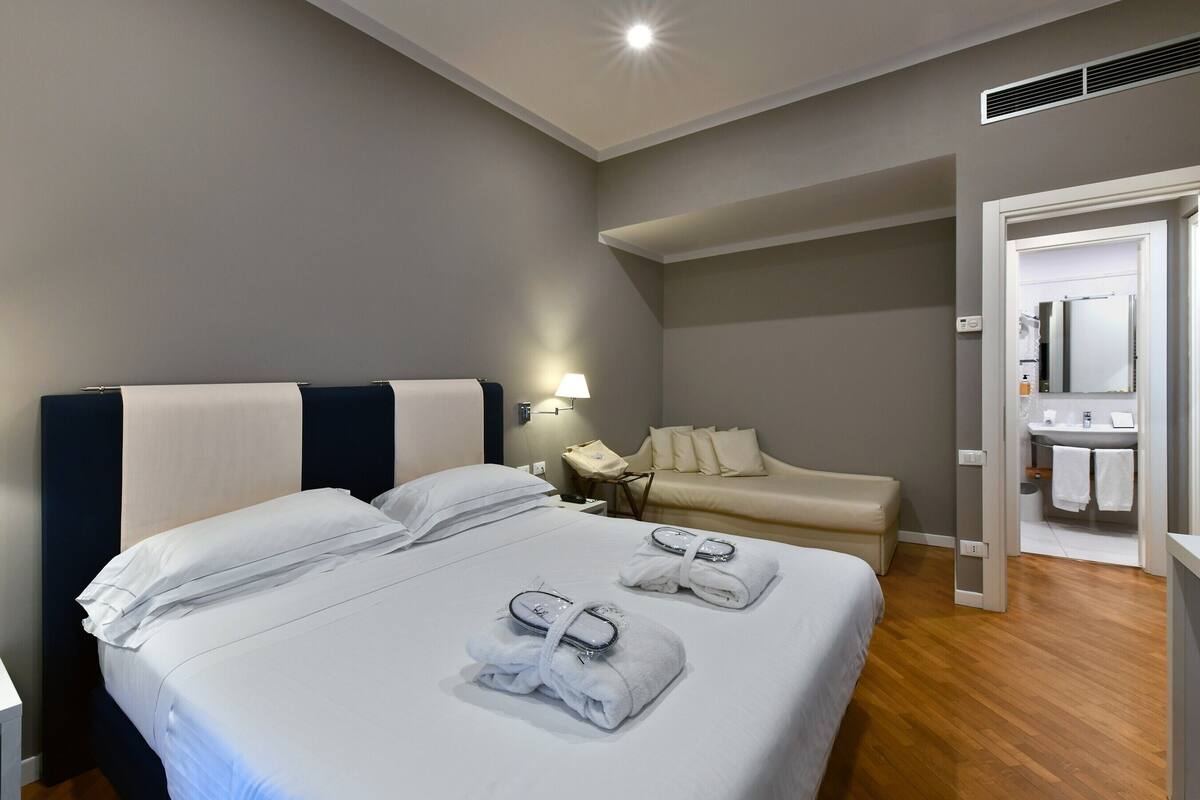 A hotel room with two beds, a sofa, a bathroom with a sink and mirror, and a door leading to another room.