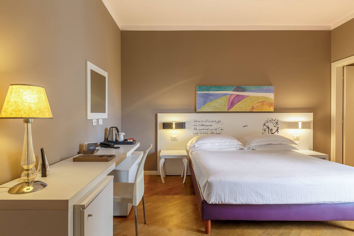 A hotel room with a bed, desk, chair, lamp, and artwork on the wall.
