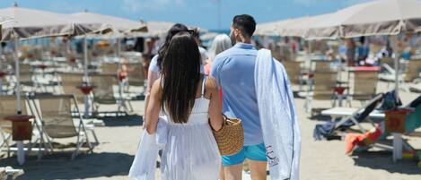 A wooden boardwalk on a sandy beach with beach chairs and umbrellas in the background.