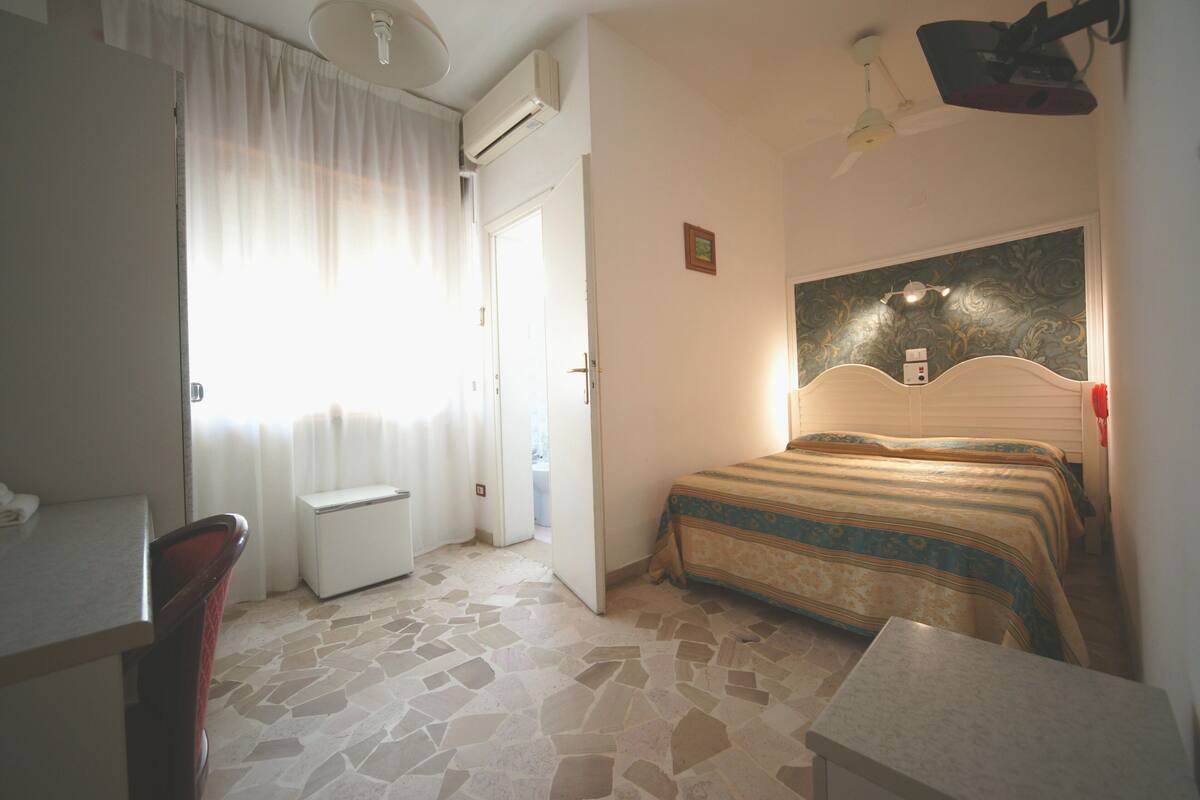 Une chambre d’hôtel avec un lit, un bureau, une chaise, une petite table et une fenêtre avec des rideaux.
