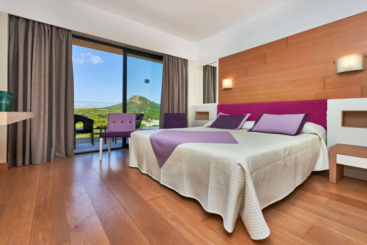 Habitación de hotel moderna con una cama grande, vistas a la montaña y un balcón con una silla.