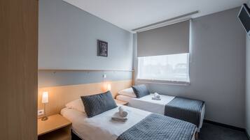 A hotel room with two beds, a bedside table with a lamp, a window with blinds, and a small picture on the wall.