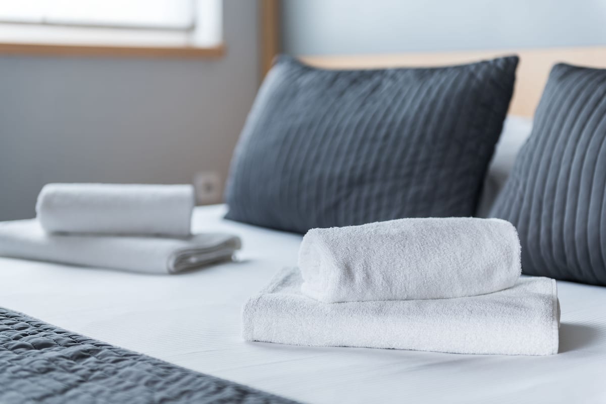 A neatly made bed with white linens and rolled towels, gray pillows, and a gray textured blanket.