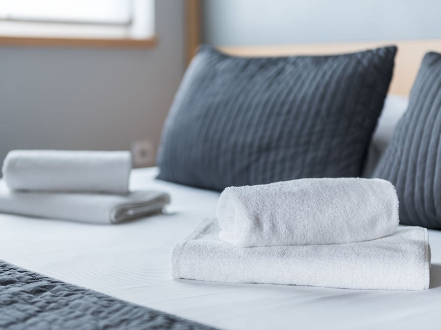 A neatly made bed with white linens and rolled towels, gray pillows, and a gray textured blanket.