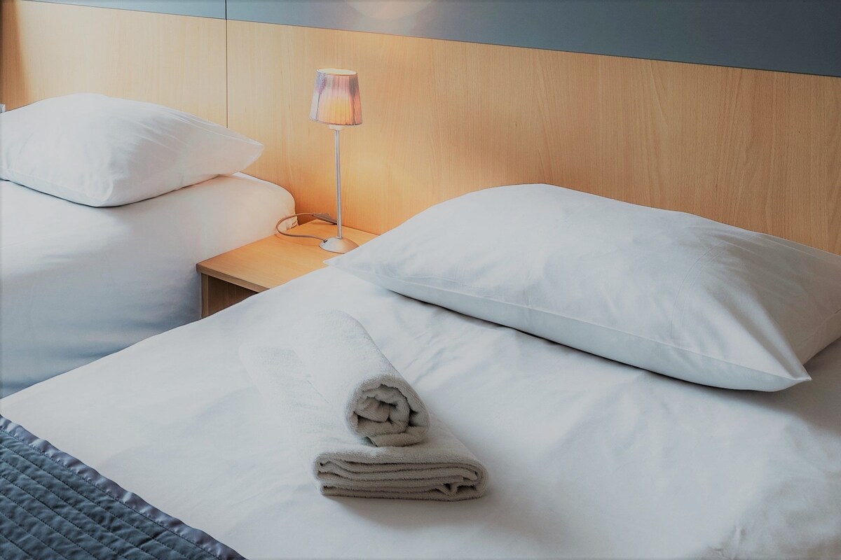 Habitació d'hotel amb dos llits, una tauleta de nit, una làmpada i una tovallola arremolinada sobre el llit.