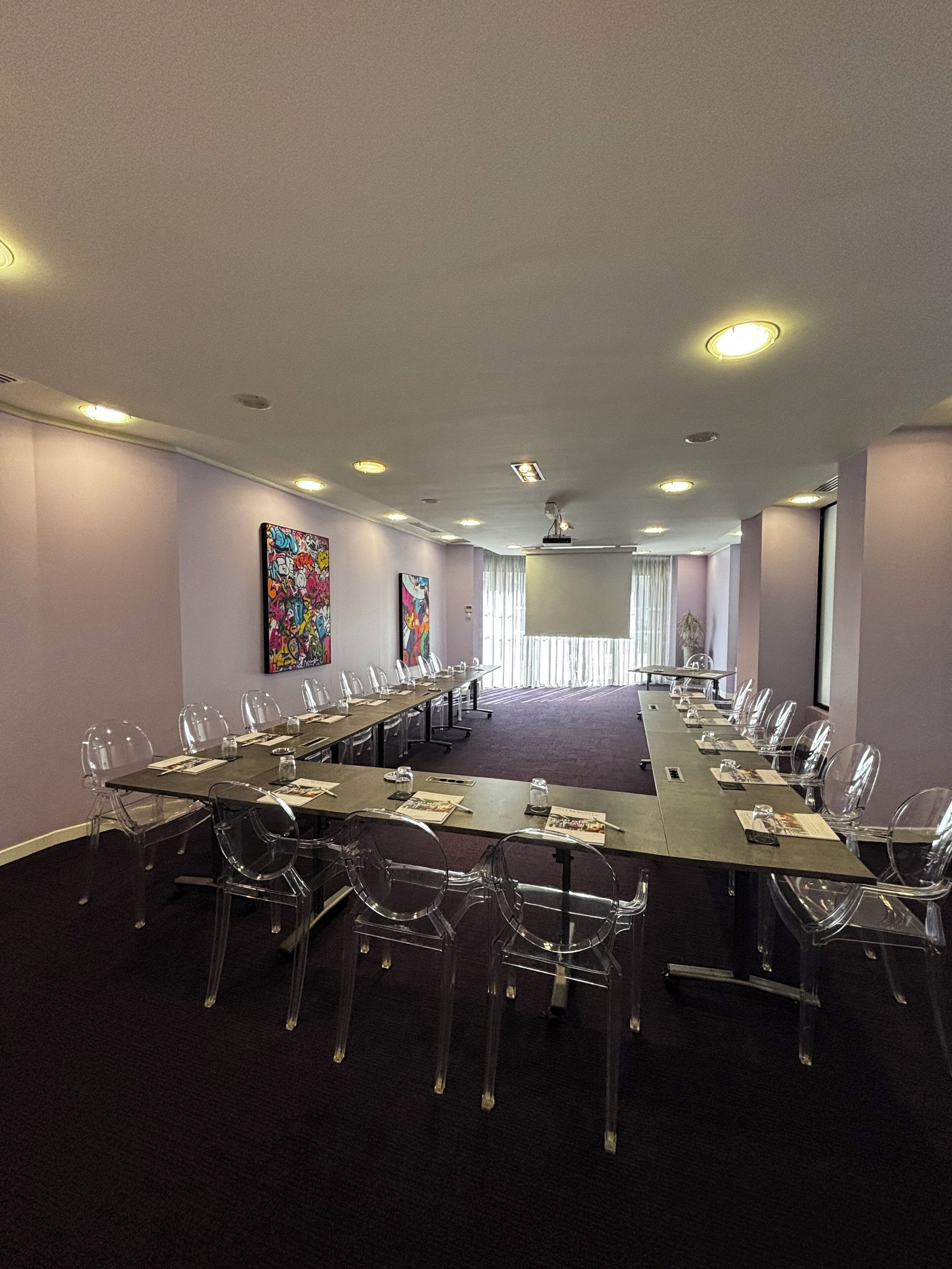 A conference room with a long table, clear plastic chairs, and ceiling lights.
