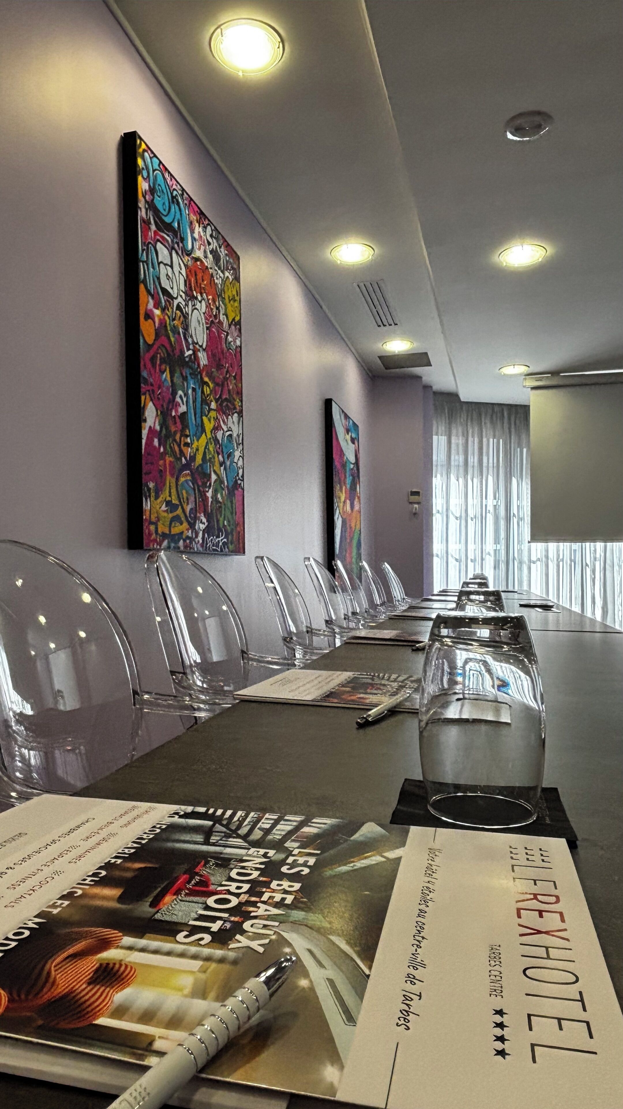 A conference room with a long table, clear plastic chairs, and two large abstract paintings on the walls.