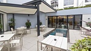 An outdoor patio with tables and chairs, a pool, and a building with large windows in the background. - Le Rex Hotel (Tarbes)