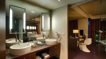 A modern hotel bathroom with two sinks, a vanity, a mirror, a hairdryer, and a shower area.