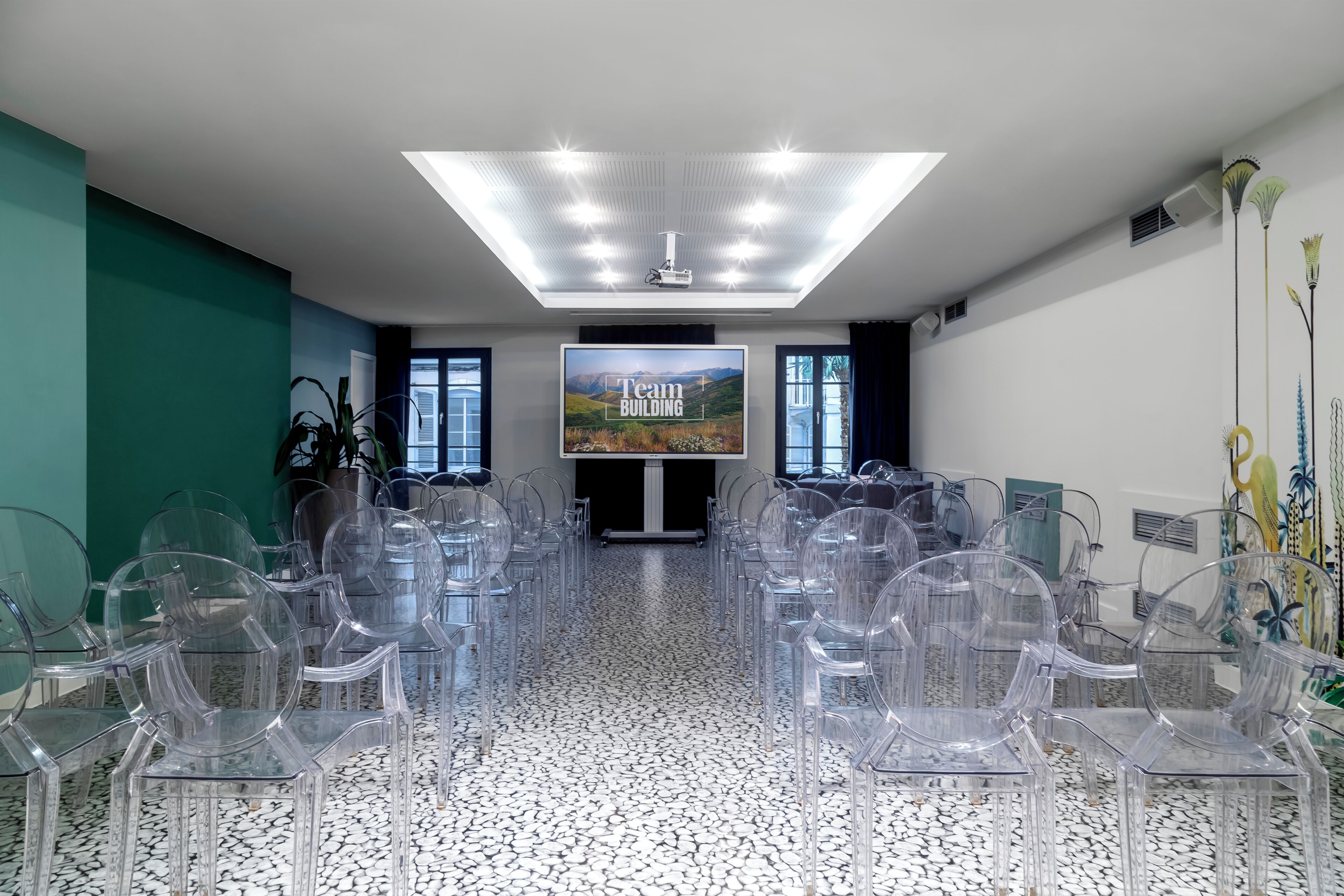 A conference room with a large screen displaying 'Team Building', clear plastic chairs arranged in rows, a ceiling with recessed lighting, and a patterned floor.