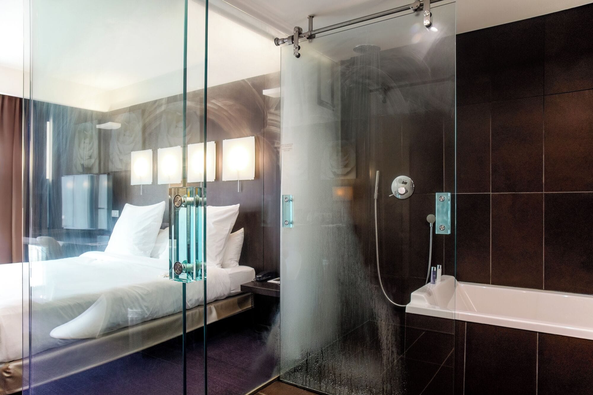 A modern hotel room with a glass partition, a bed, a shower area, and a bathtub.