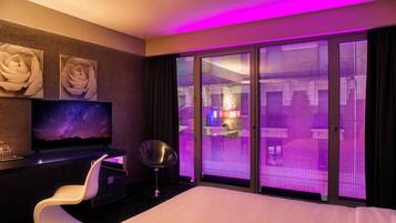 A hotel room with a bed, a desk with a computer, a chair, and a large window with a view of the city.