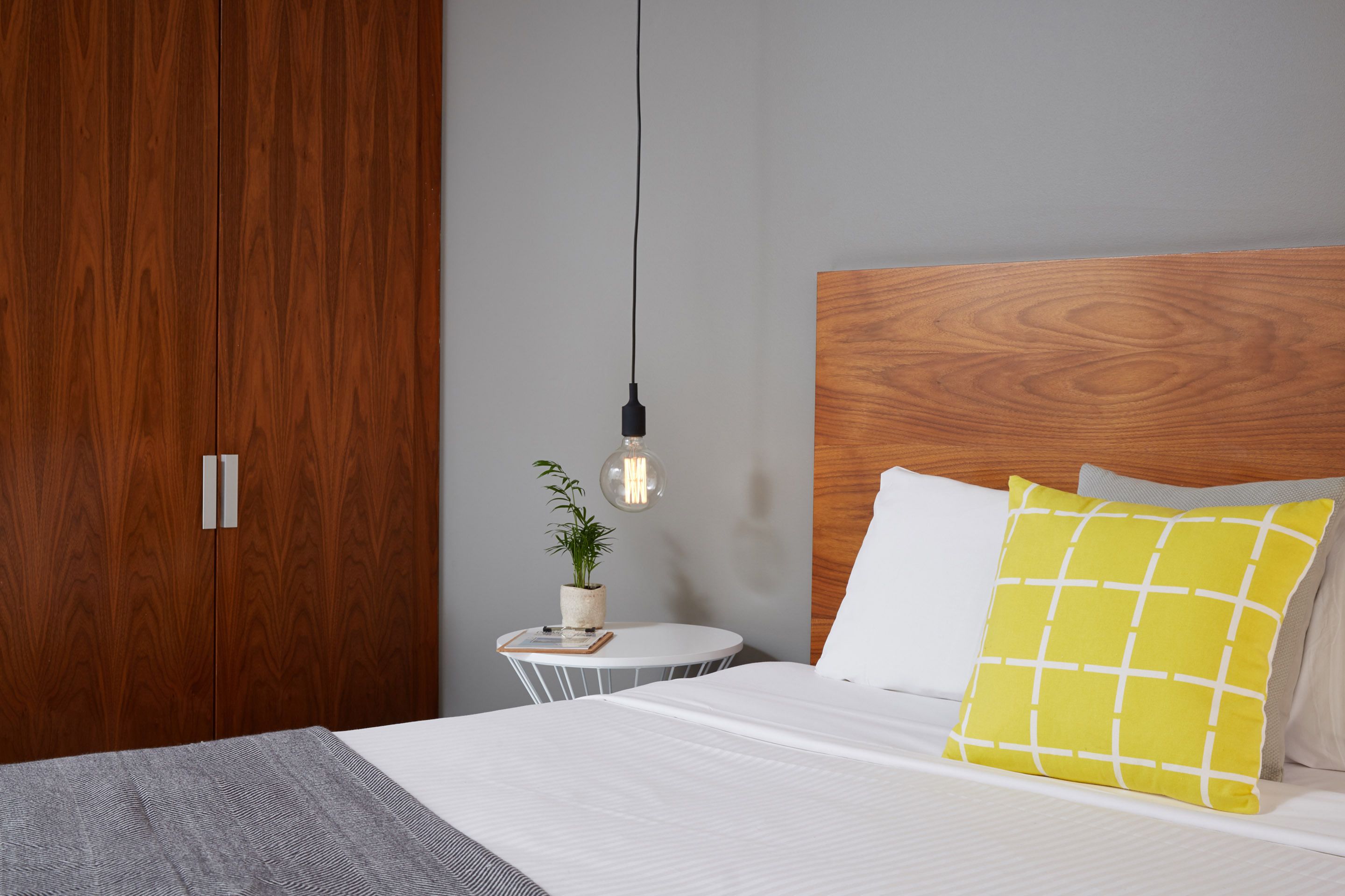 Ein ordentlich bezogenes Bett mit einem weißen und gelben Kopfkissen, ein Nachttisch mit einer Pflanze und ein Holzschrank.