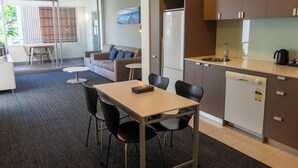 A modern kitchen with a dishwasher, a dining area with a table and chairs, and a sofa in a living area with a view of the outdoors. - Cosmopolitan Hotel and Apartments (St Kilda)