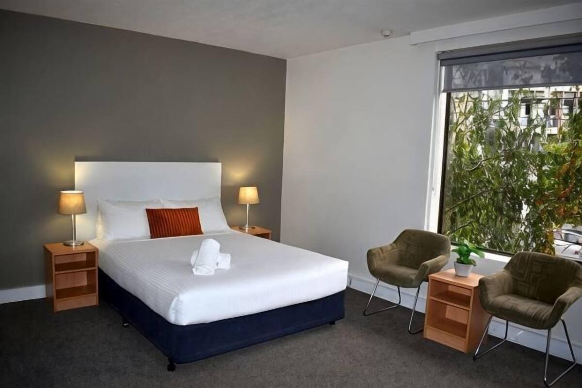 Une chambre d’hôtel avec un grand lit, deux tables de chevet, une chaise, une petite table avec une plante et une fenêtre donnant sur des arbres.