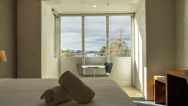 A hotel room with a bed, a table, and chairs near a large window with a city view. - Cosmopolitan Hotel and Apartments (St Kilda)