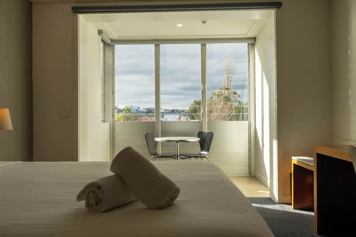Quarto de hotel com cama, mesa e cadeiras perto de uma janela grande com vista para a cidade.