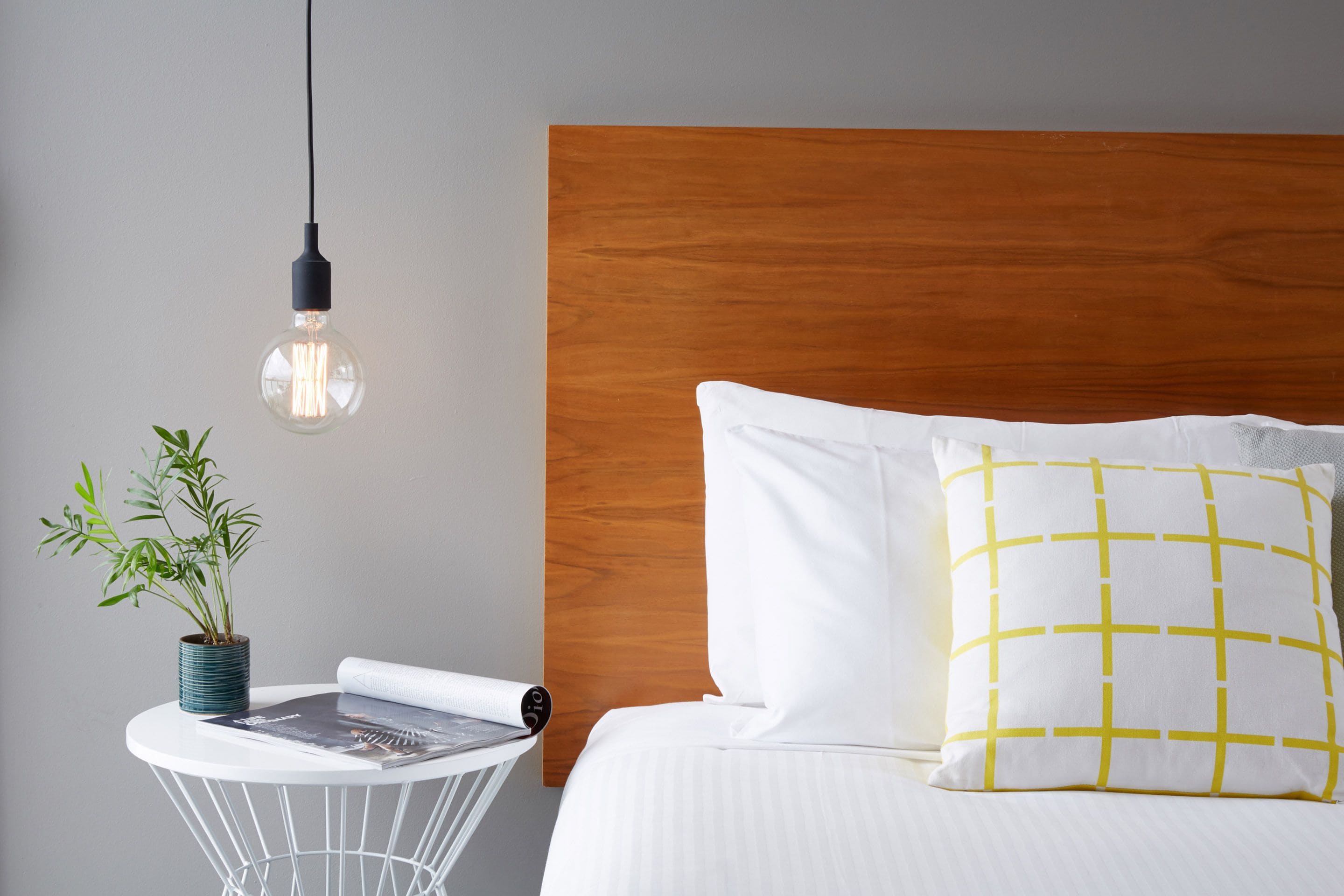 A neatly made bed with white and yellow pillows, a wooden headboard, a small round table with a plant and a magazine, and a hanging light bulb.