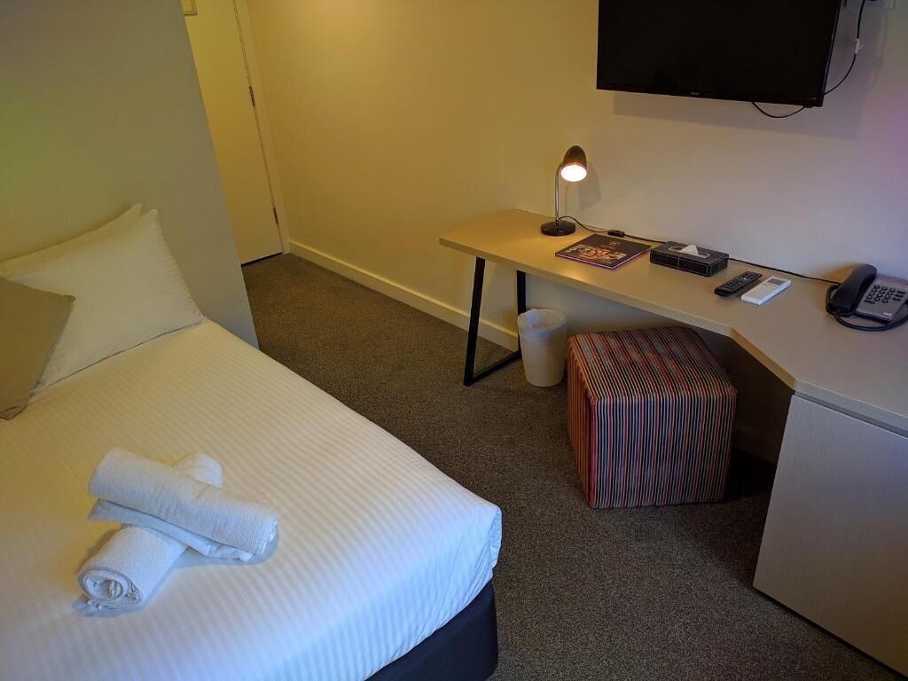 Quarto de hotel com cama, escrivaninha com luminária, telefone e televisão na parede.