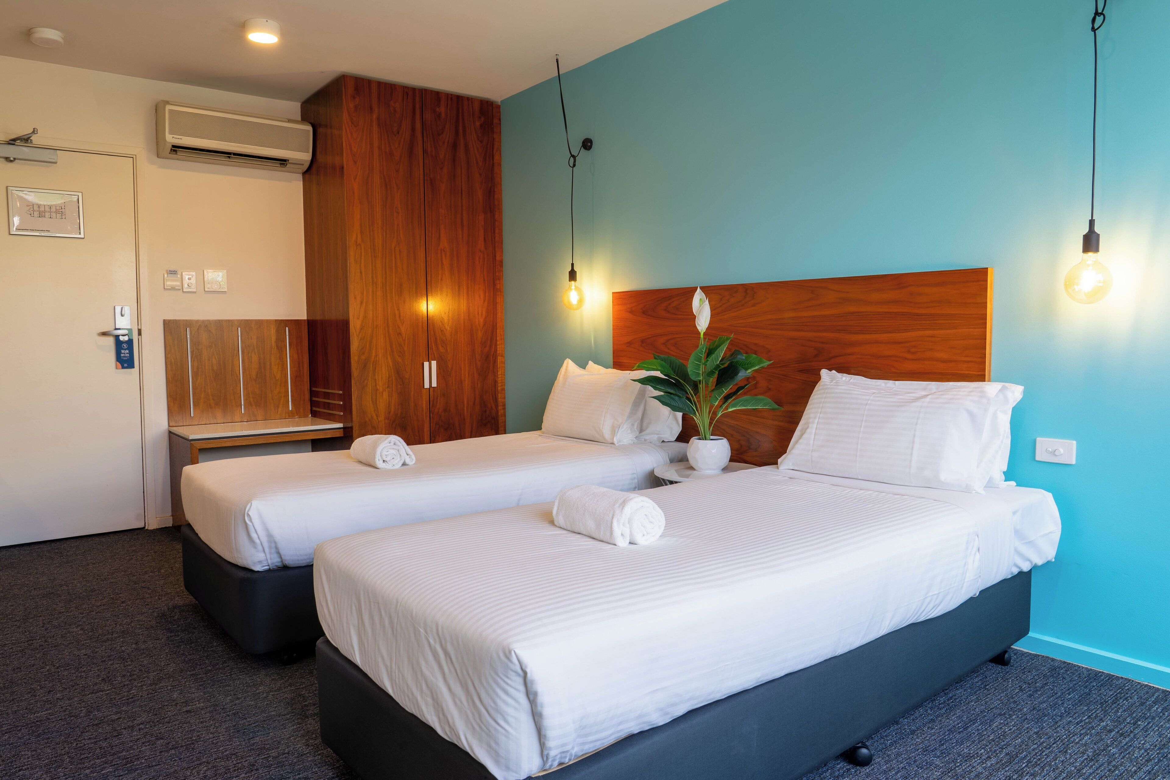 Een hotelkamer met twee bedden, een houten garderobe, een potplant en een airconditioner aan de muur.