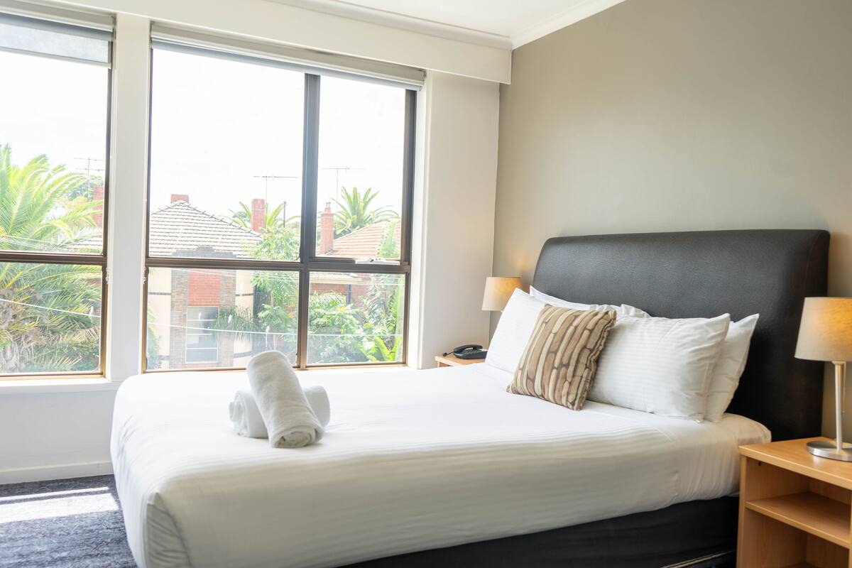 Quarto de hotel com uma cama grande, duas luminárias de cabeceira e uma janela com vista para edifícios e áreas verdes.