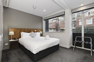 Una habitación de hotel moderna con una cama grande, una mesita de noche con lámpara, una ventana con vistas a edificios y un balcón con barandilla de metal.