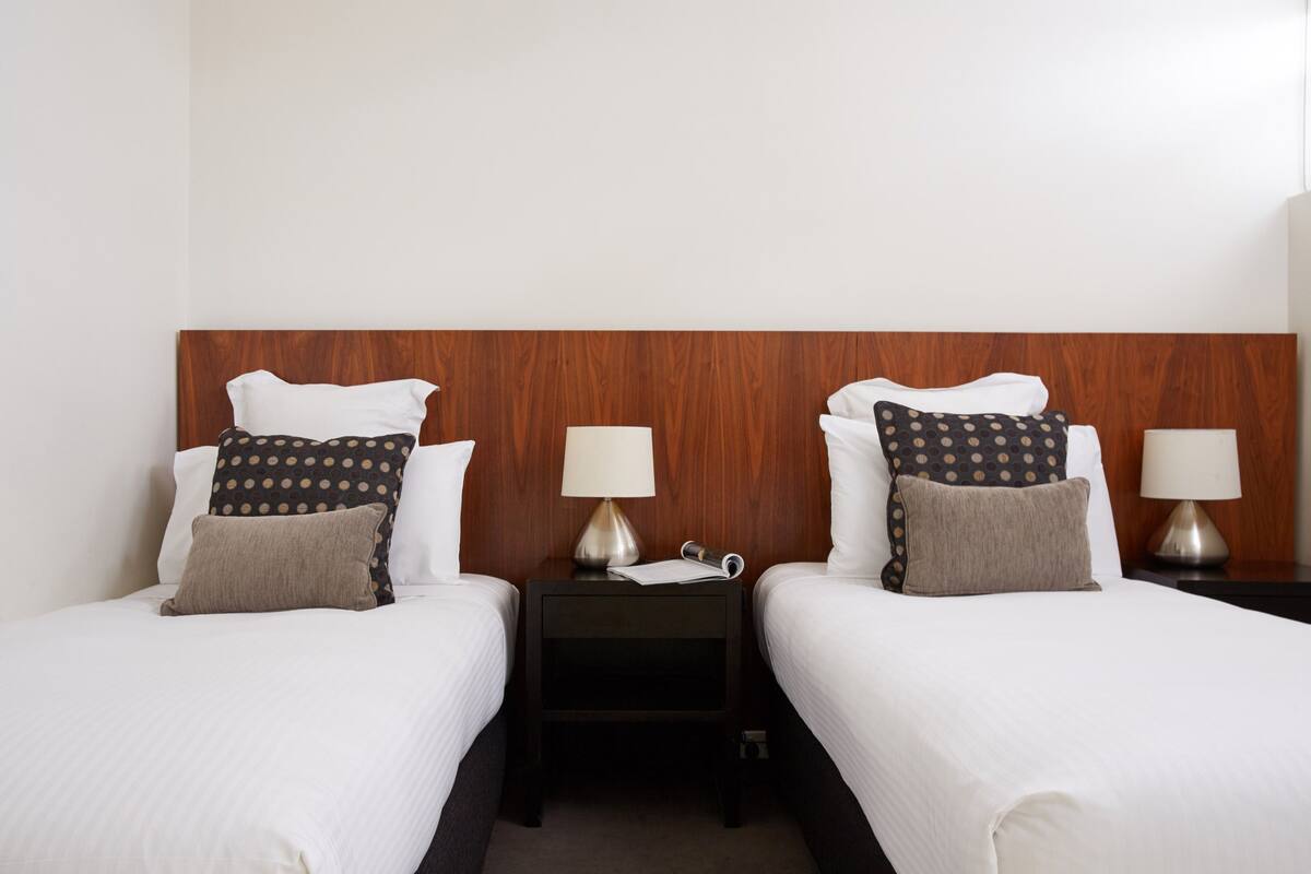 Quarto de hotel com duas camas de solteiro, cada uma com travesseiro e mesa de cabeceira com abajur.