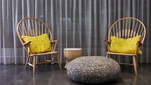 Two wooden chairs with yellow cushions, a round wooden side table, and a grey knitted ottoman in a room with grey curtains. - Cosmopolitan Hotel and Apartments (St Kilda)
