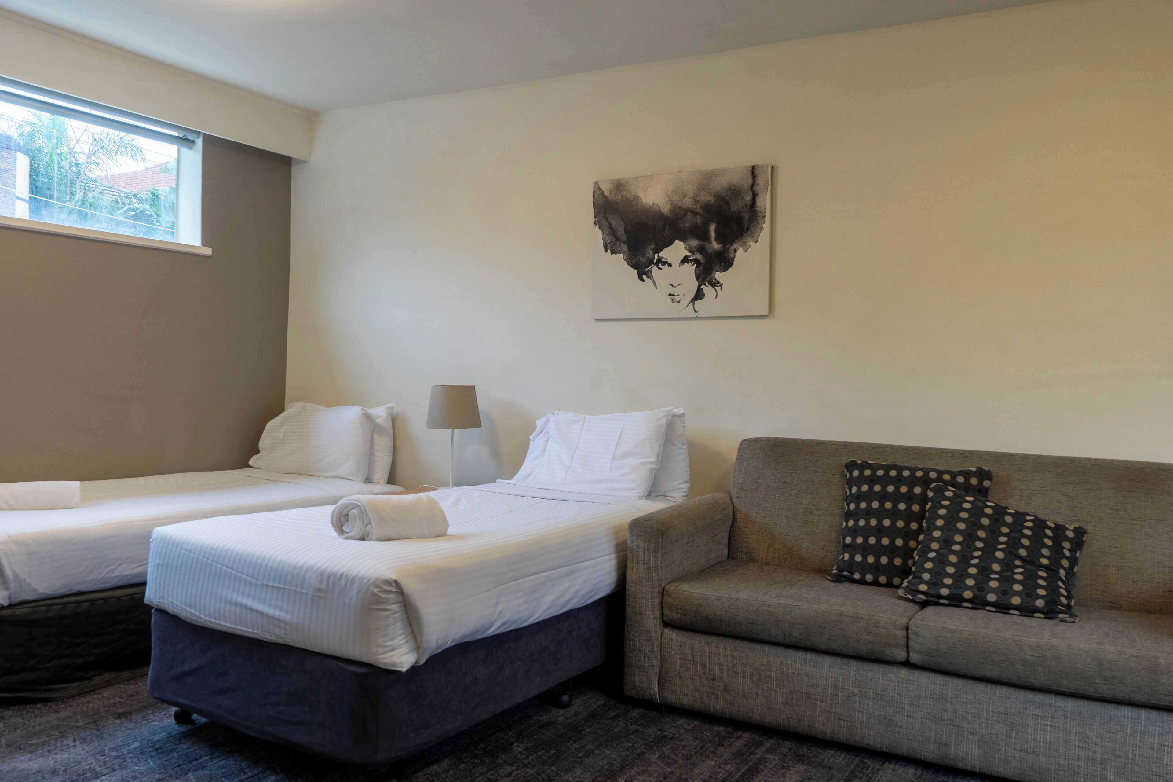 A hotel room with two single beds, a sofa, a lamp, and a painting on the wall.