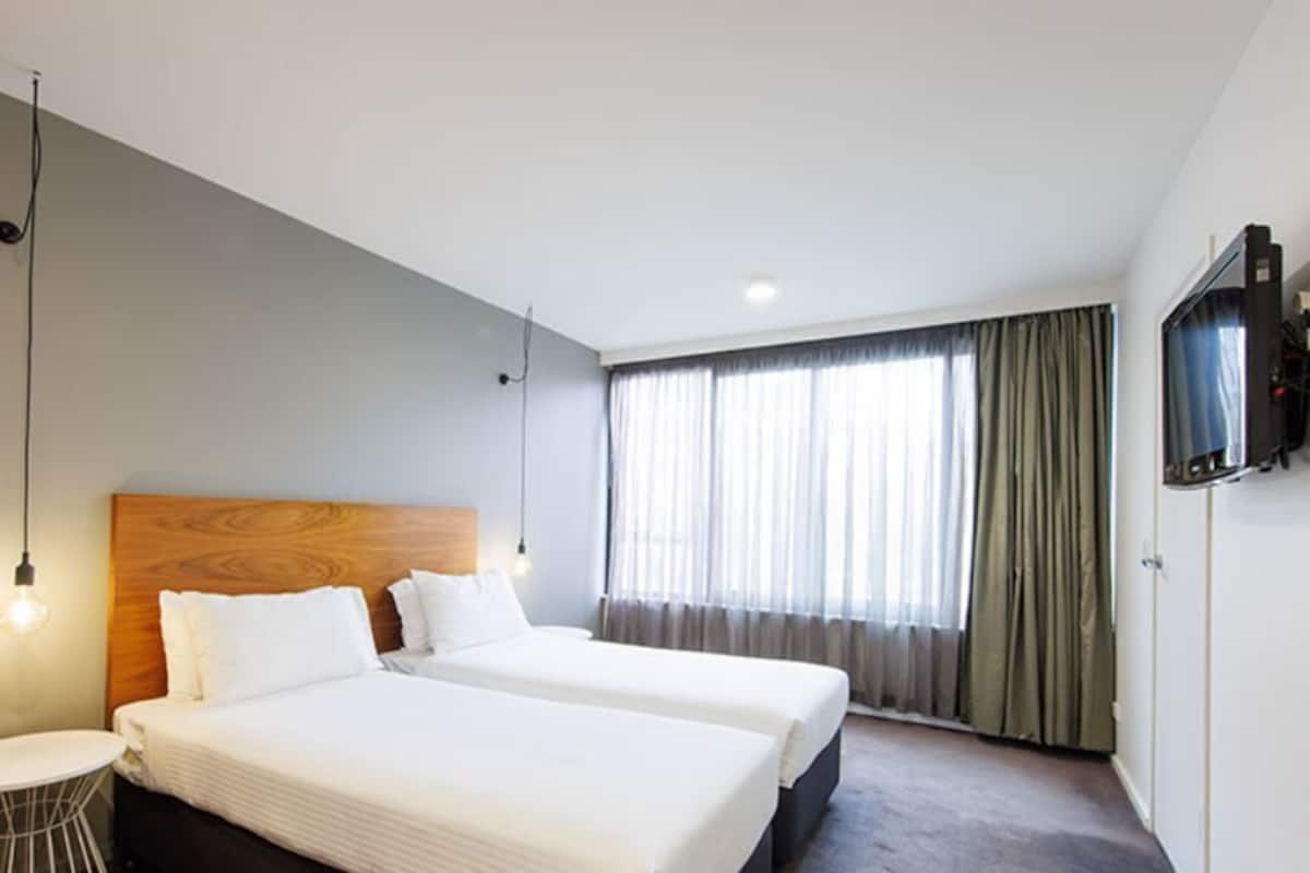 Quarto de hotel com duas camas, uma televisão, uma mesa redonda e uma janela com cortinas.
