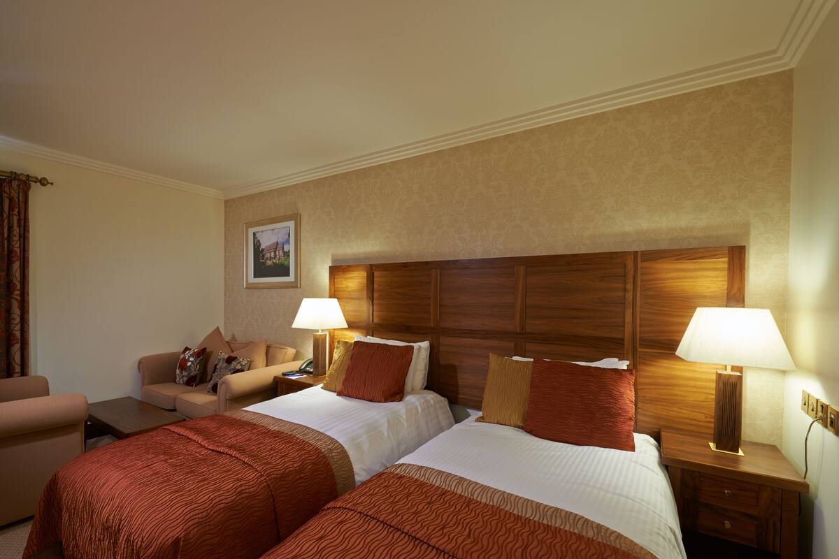 Quarto de hotel com duas camas, um sofá, uma escrivaninha e uma luminária.