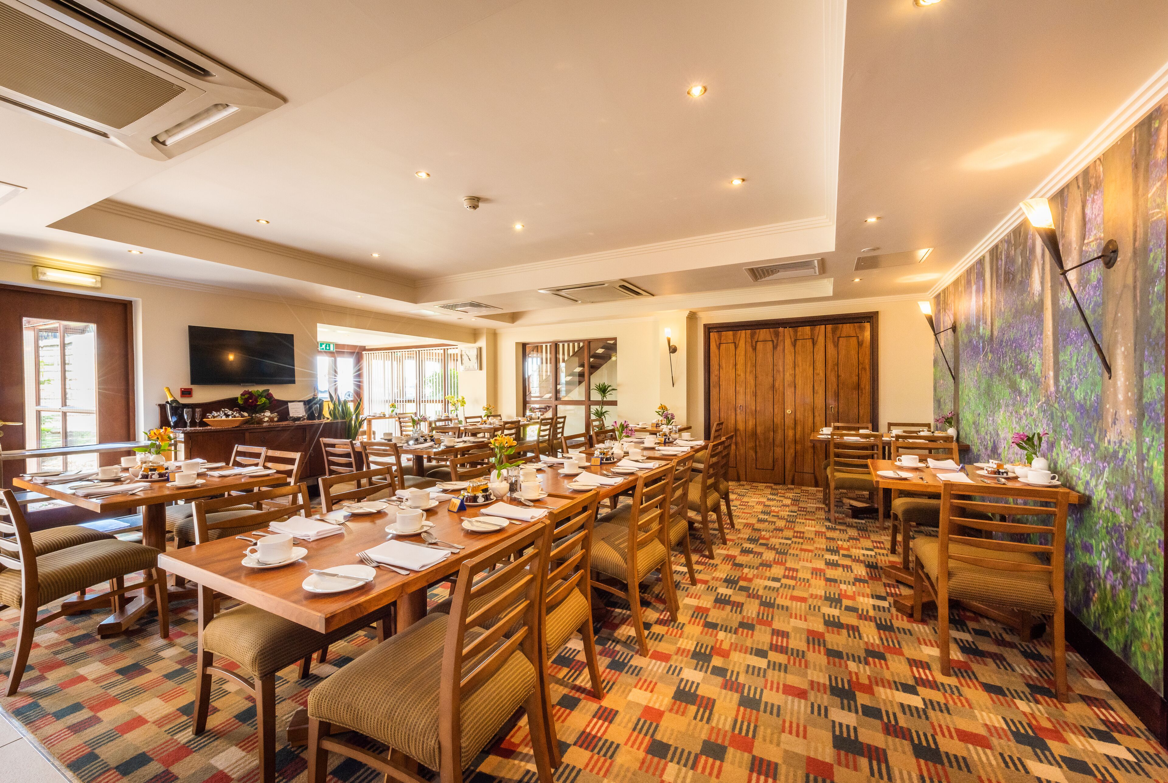 A dining area with multiple tables set for meals, a television mounted on the wall, and a patterned carpet.