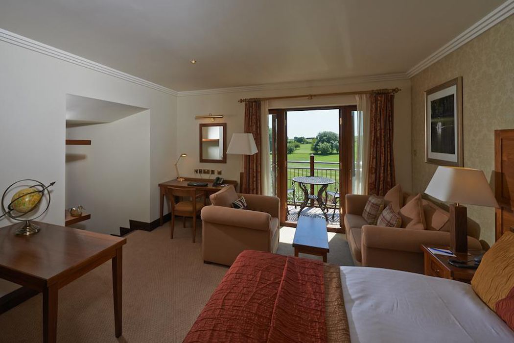 A hotel room with a bed, two armchairs, a small table, a desk, and a balcony with a view of the garden.