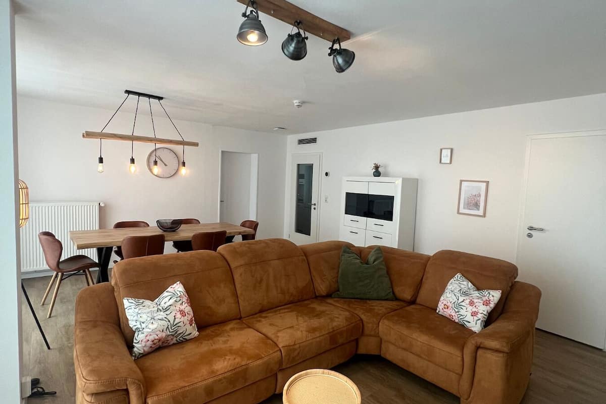 A modern living room with a brown sectional sofa, a small round table, and a dining area with a wooden table and chairs.