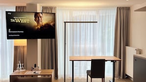 Ein modernes Hotelzimmer mit einem Flachbildfernseher, der 'Kevin Costners Der Westen' zeigt, einem Holztisch, einem Sessel, einem Nachttisch mit Lampe und einem großen Fenster mit Vorhängen.