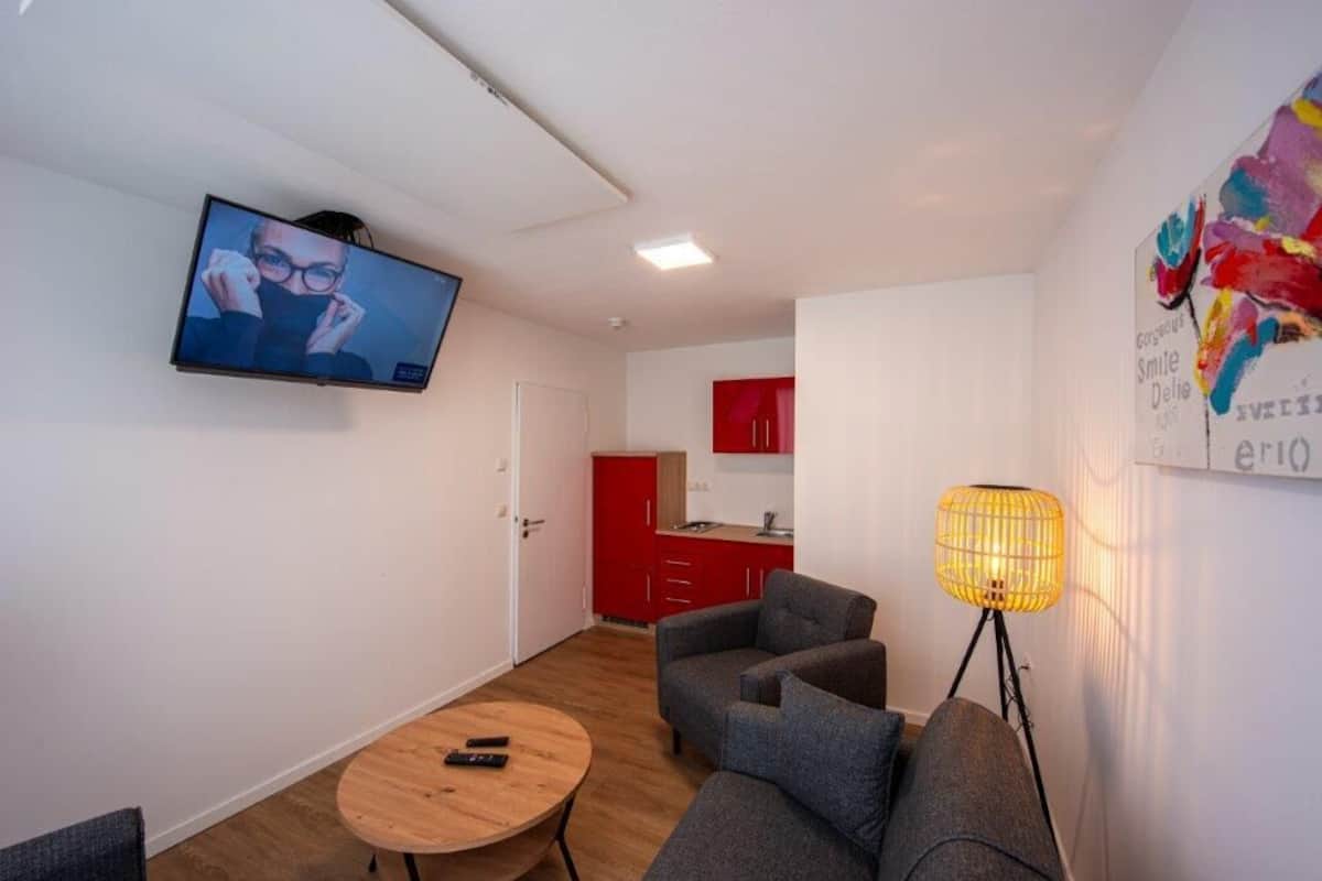 A modern living room with a TV, grey sofas, a round wooden table, and a red kitchen in the background.