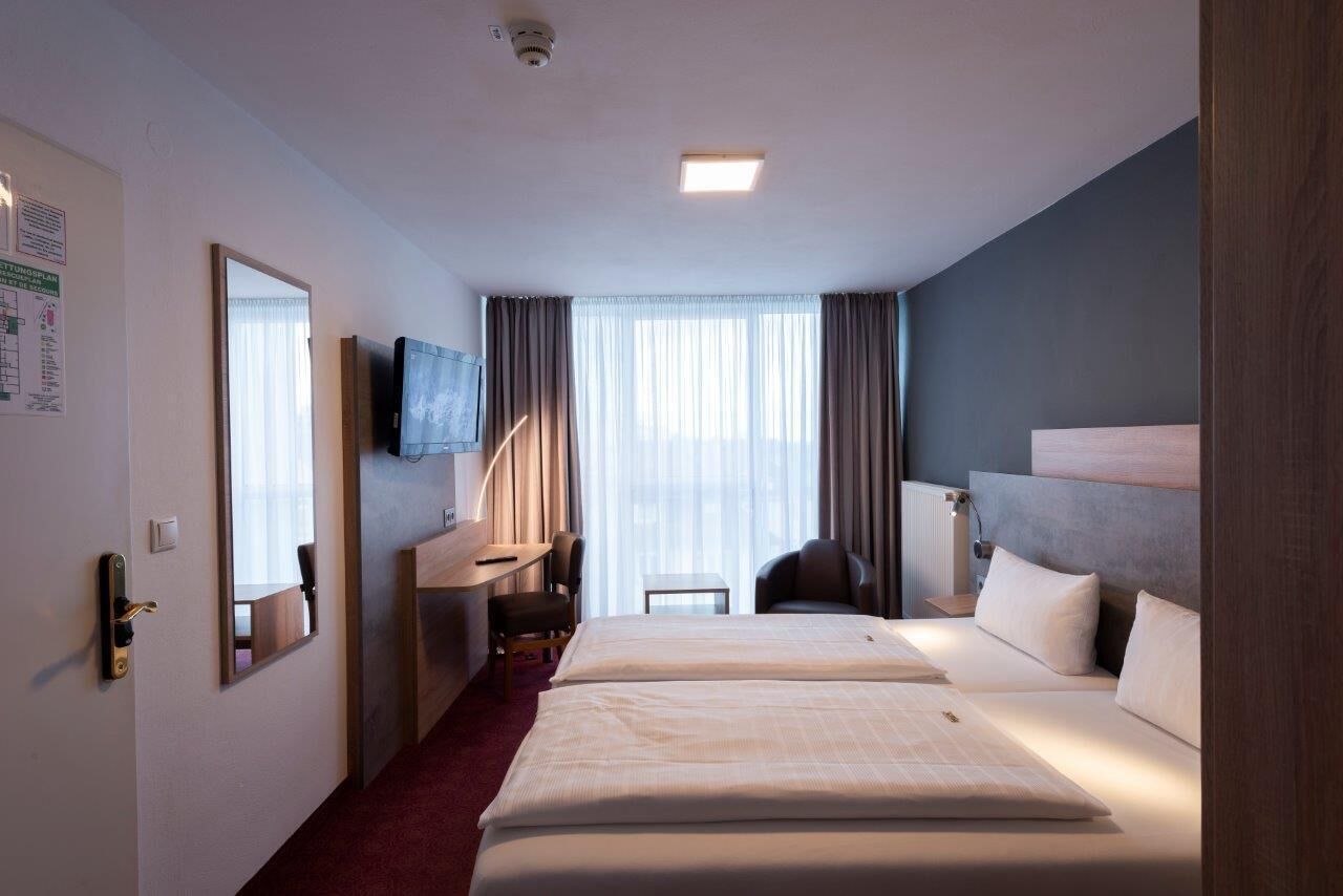 Ein Hotelzimmer mit zwei Betten, einem Schreibtisch, einem Stuhl, einem Fernseher und einem Fenster mit Vorhängen.