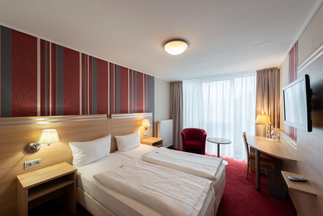 Habitación de hotel con dos camas, un escritorio, una silla, un televisor y una ventana con cortinas.
