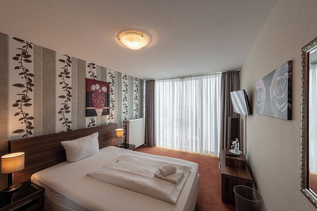 A hotel room with a large bed, a bedside table with a lamp, a TV mounted on the wall, and a window with curtains.