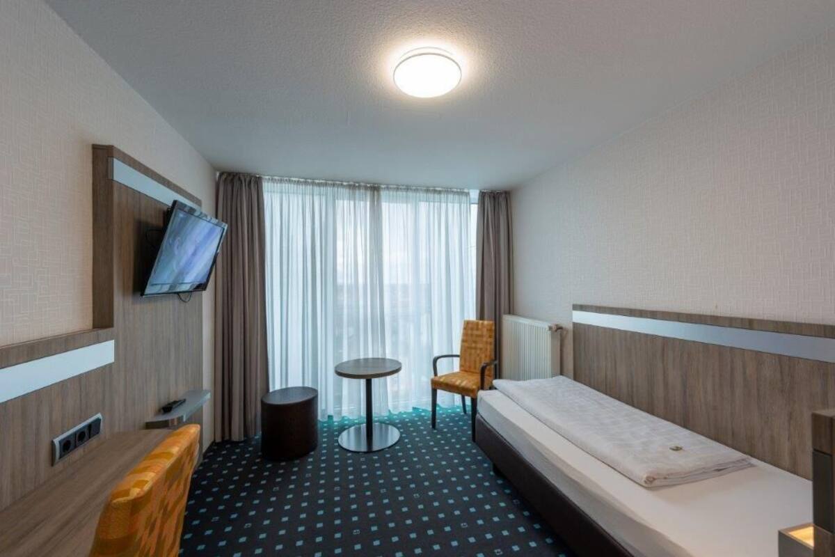 A hotel room with a bed, a chair, a small table, a television, and a window with curtains.