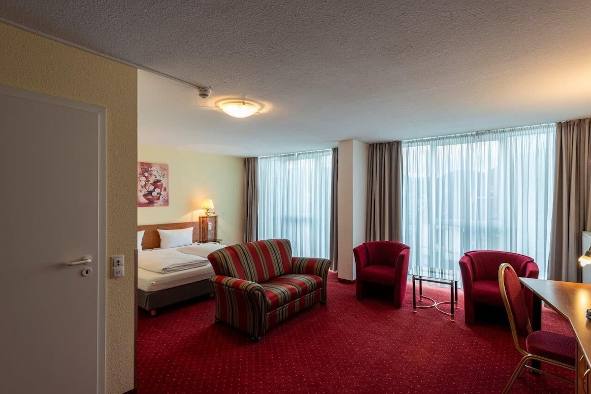A hotel room with a bed, a striped sofa, two red armchairs, a small table, and a window with curtains.