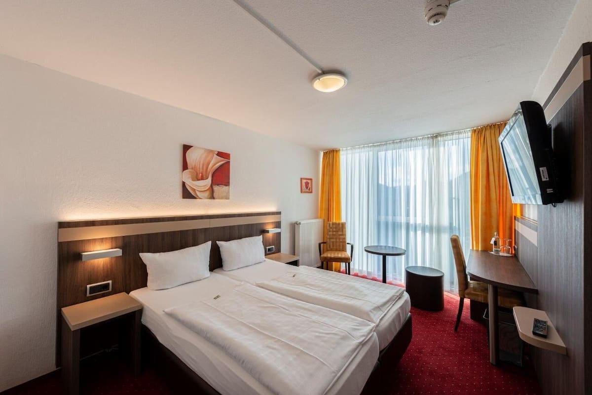 A hotel room with a large bed, bedside tables, a desk, a chair, a TV, and a window with curtains.