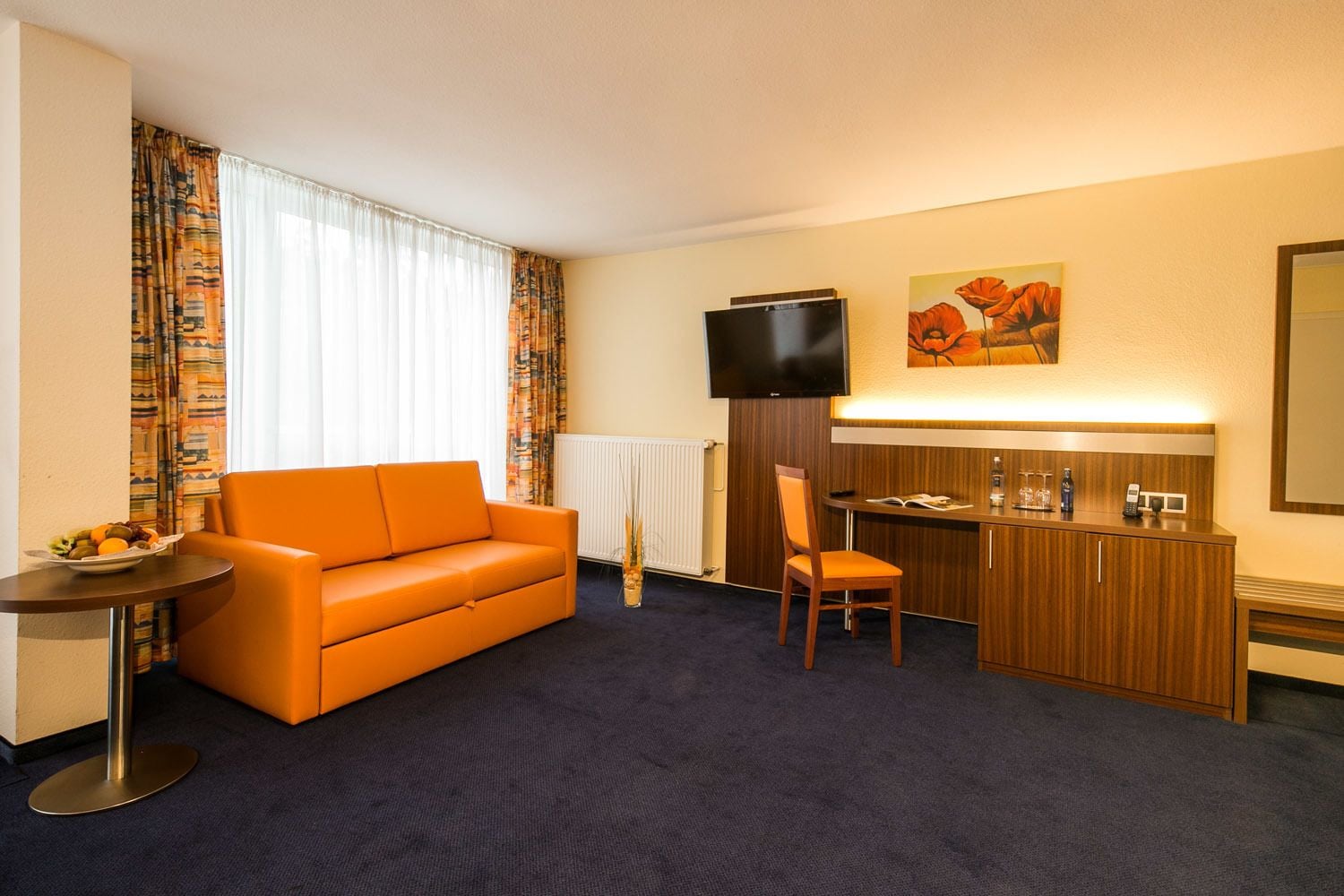 A hotel room with a sofa, a small table with fruit, a desk with a chair, a TV, and a painting on the wall.