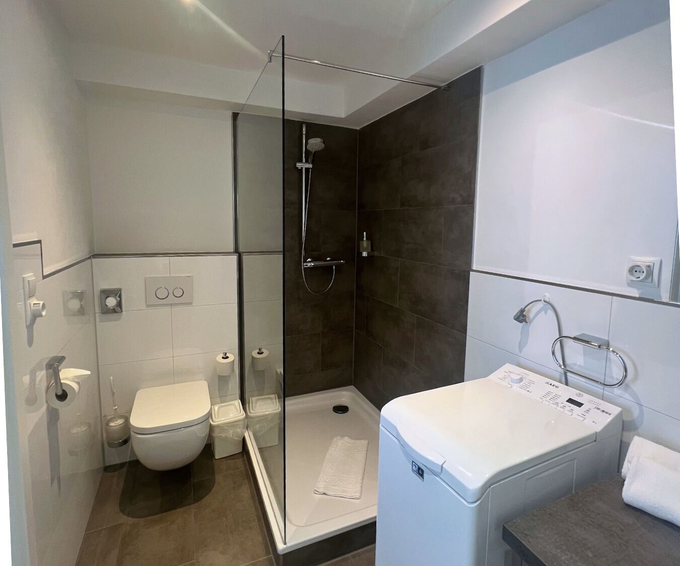 A modern bathroom with a glass-enclosed shower, a toilet, a washing machine, and a towel rack.
